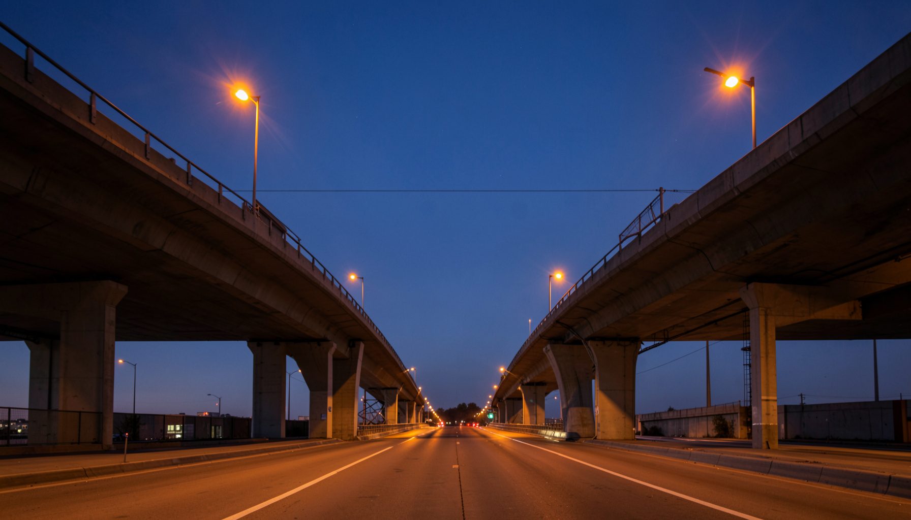 A clean, symmetrical architectural photograph of a modern highway overpass at dusk, with warm amber streetlights reflecting off concrete and steel, deep blue sky above